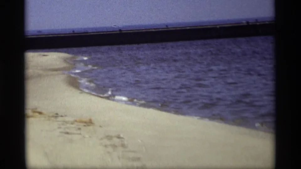 A 1969 8mm home-movie view of the Holland, Michigan shoreline, with rippled water and pale sand framed by dark film edges.