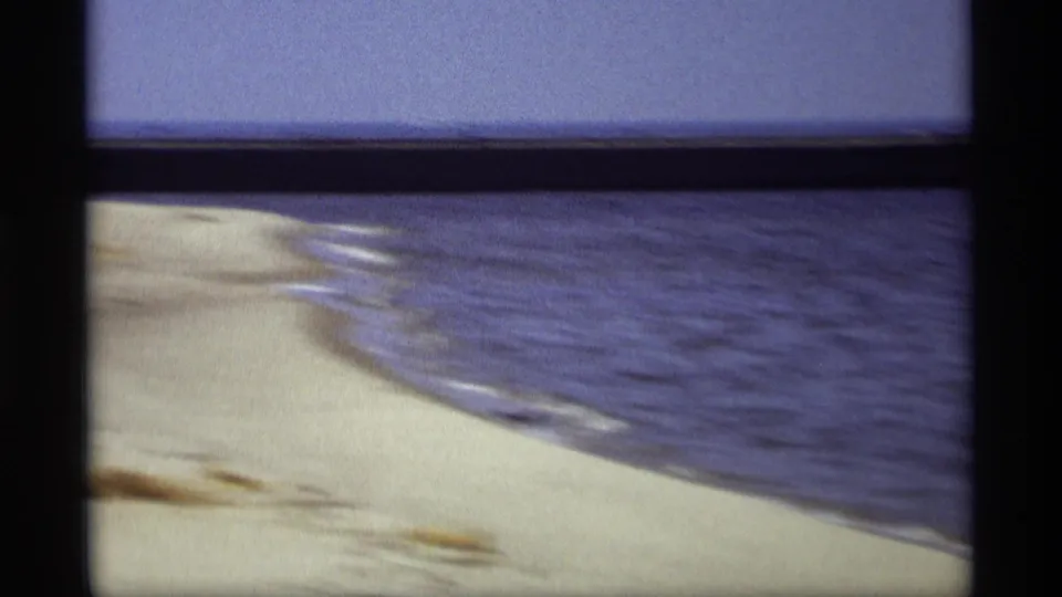 The lakeshore curves through the frame, showing pale sand, blue water, and faint wave lines in soft 8mm color.