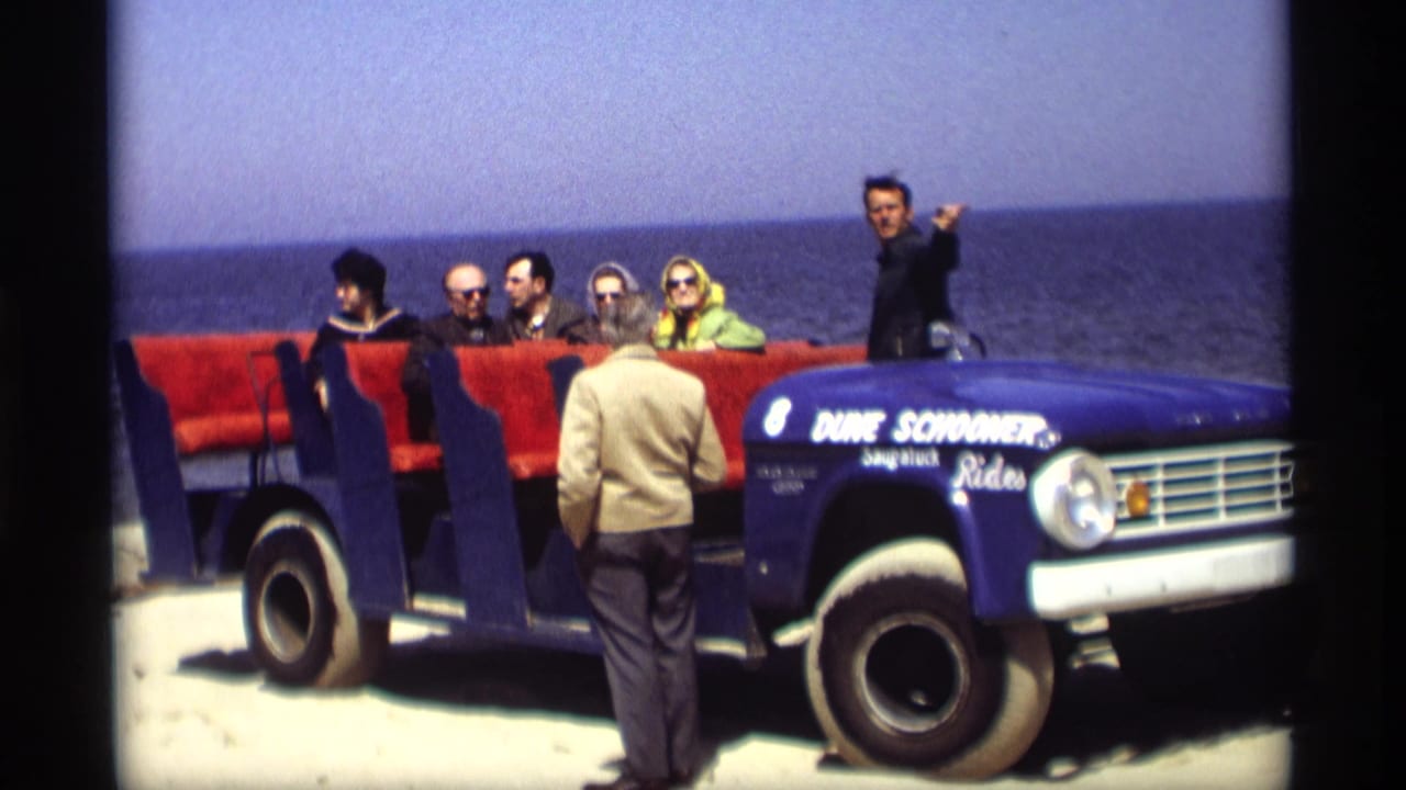 Tourists seated in a blue open tour vehicle beside water as a guide stands near the front.