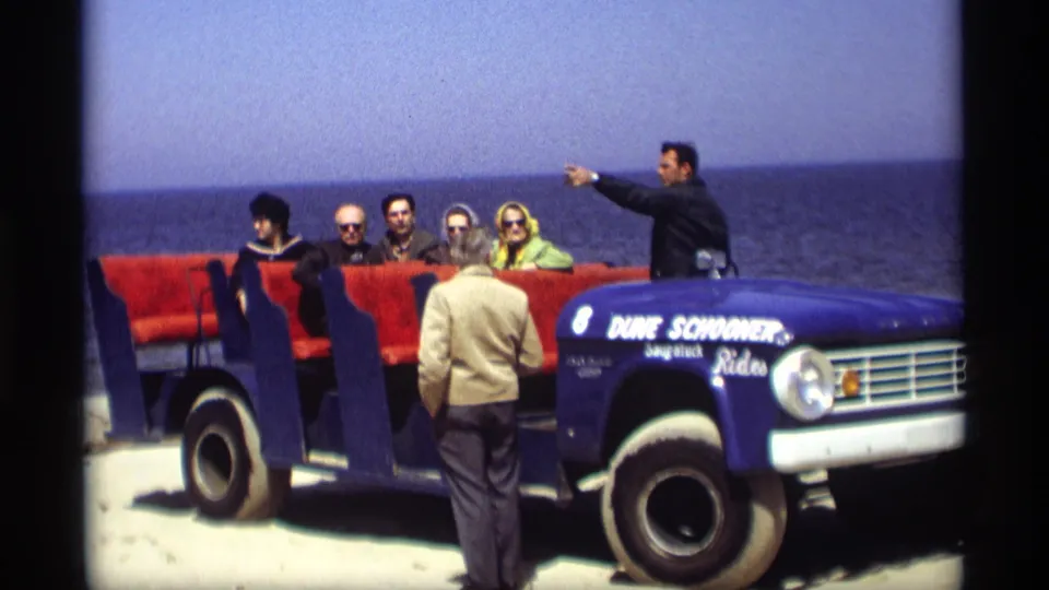 A guide points beside a blue Dune Schooner Rides vehicle carrying adult passengers along a Holland, Michigan shoreline in 1969.