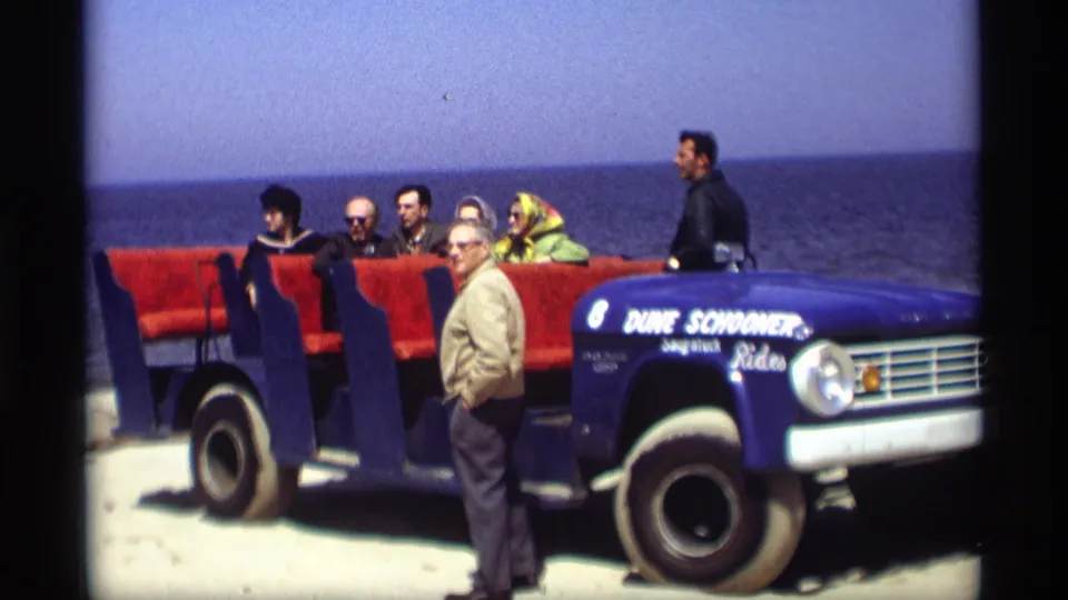 An older man stands by the blue Dune Schooner Rides vehicle as adult tourists sit behind him near the Holland, Michigan shoreline.