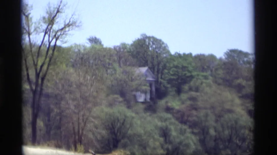 1969 Holland, Michigan 8mm frame showing a distant white house nestled in dense trees beyond a dark vertical frame edge.