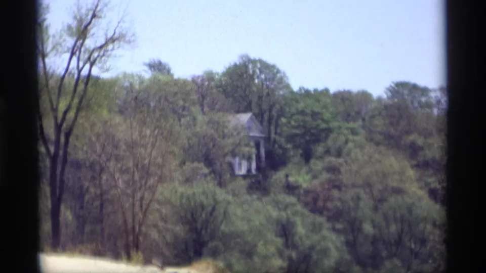1969 Holland, Michigan 8mm view of a partially obscured house across a wooded slope, framed by dark vertical borders.