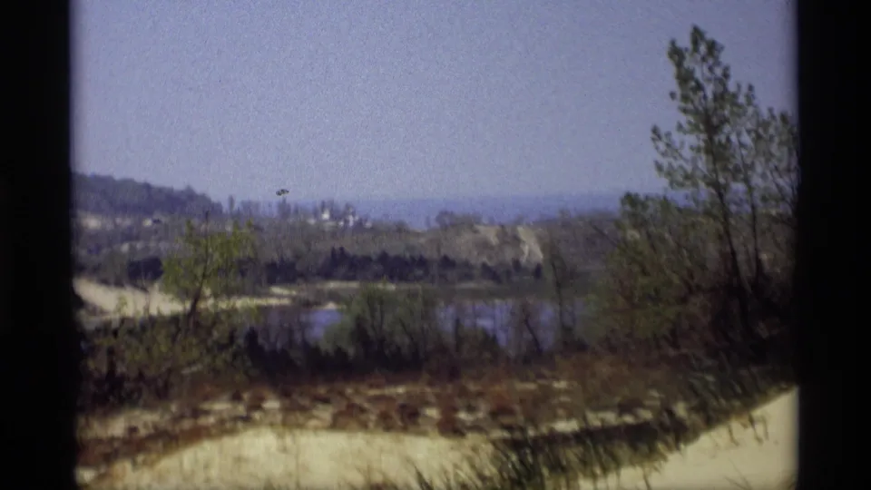 1969 Holland, Michigan 8mm frame with a calm pond, brushy shoreline, sandy banks, trees, and hazy low hills in the distance.