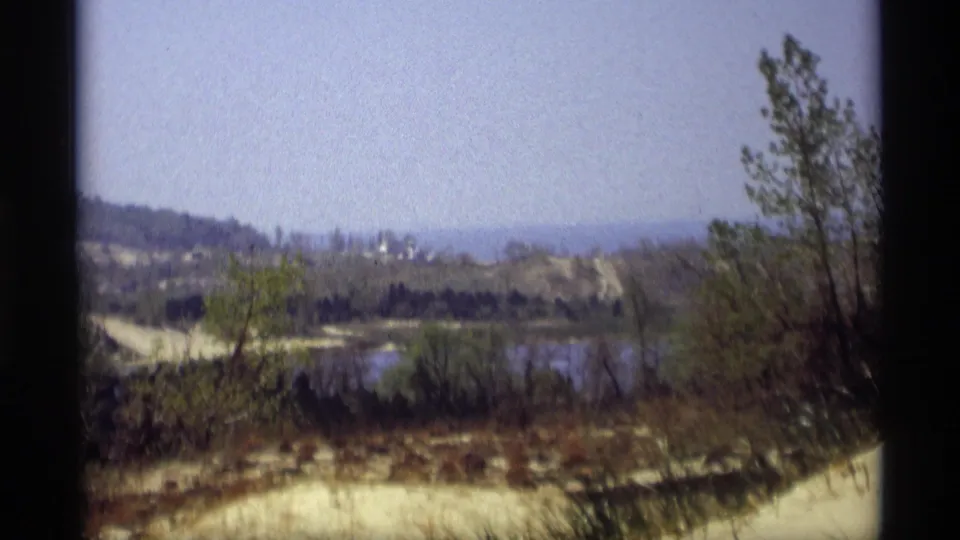 1969 Holland, Michigan 8mm frame looking across a pond and brush toward sandy banks, trees, and hazy hills beyond.