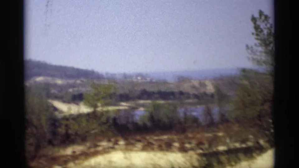 1969 Holland, Michigan 8mm frame with soft-focus pond water, scrub vegetation, exposed sand, and a distant wooded ridge.