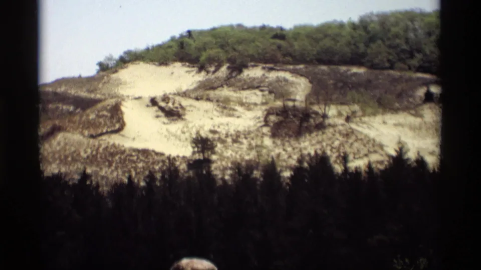 1969 Holland, Michigan home-movie frame showing exposed pale soil and blackened ground below a green wooded ridge.