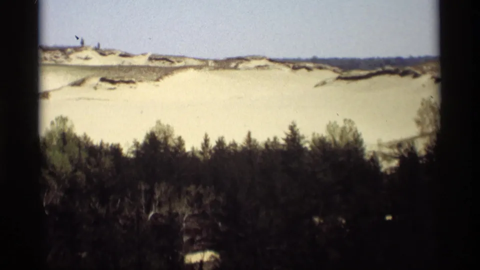 Wide 1969 8mm view of Holland, Michigan dunes, a dark treeline, and tiny distant people on the sand ridge.