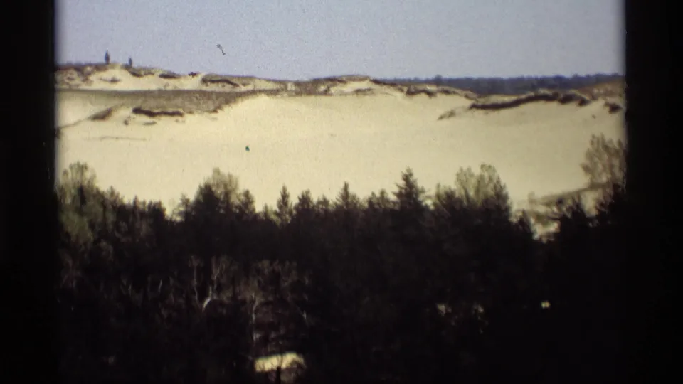 1969 8mm Holland, Michigan view of a broad pale dune, dark pine forest, and small distant visitors near the horizon.