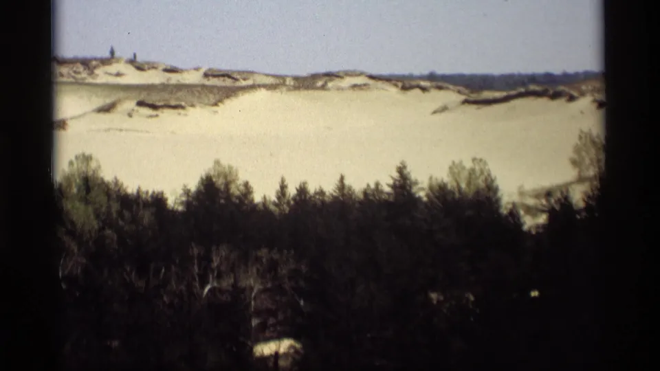 1969 8mm frame from Holland, Michigan showing snowy dunes rising behind a dense line of winter trees.