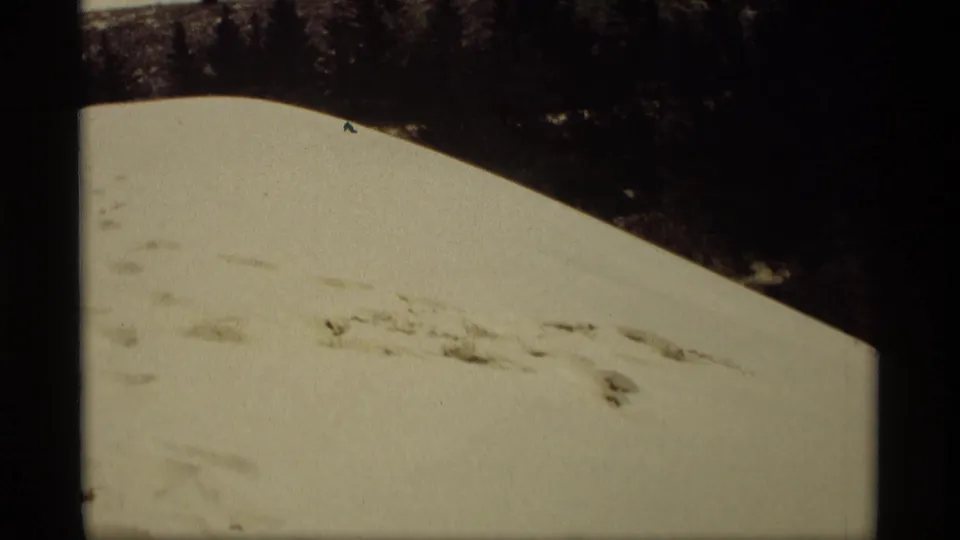 1969 Holland, Michigan winter frame with a pale snowbank, scattered surface marks, and dark trees beyond the hill crest.