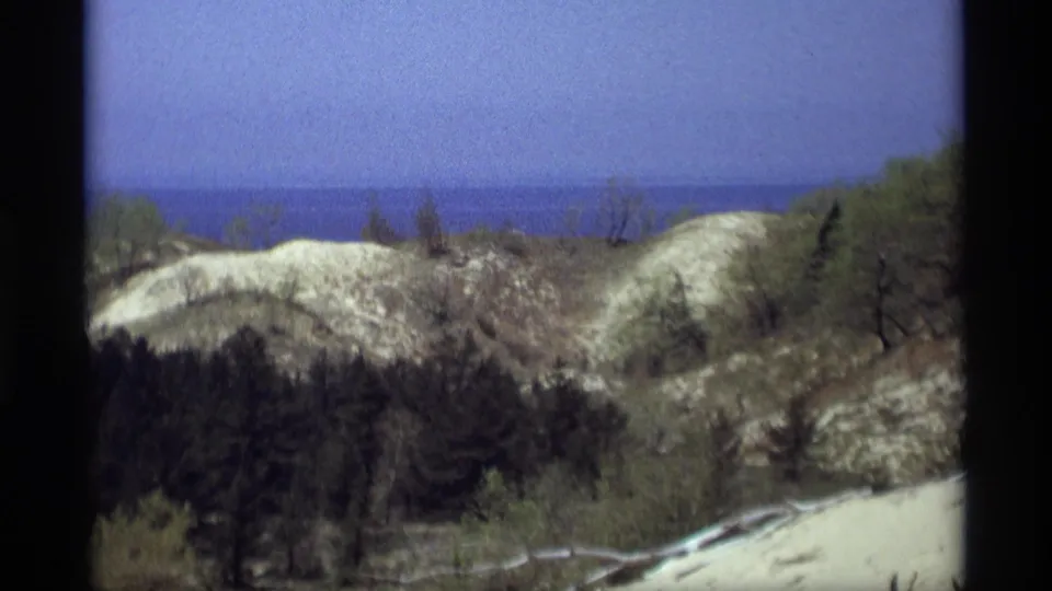 A wider 1969 Holland, Michigan view shows rolling pale dunes, darker evergreens, brush, and distant Lake Michigan water beyond the ridge.