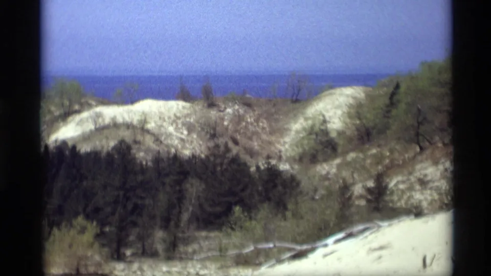 A wide home-movie frame shows pale dune ridges, scattered vegetation, dark trees, and blue water on the horizon near Holland, Michigan.