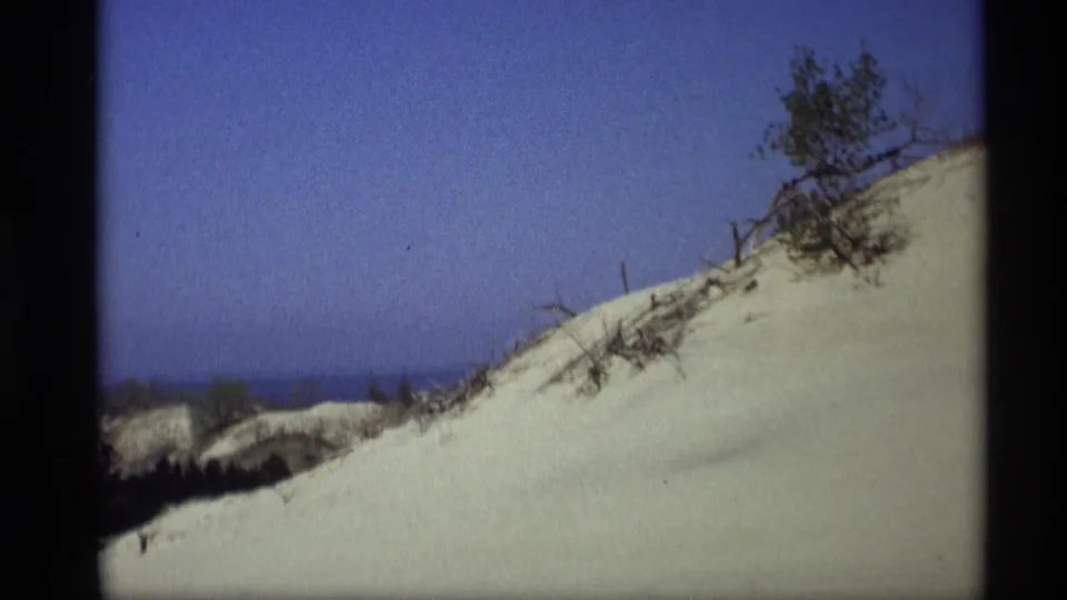 Vintage 8mm frame of Holland, Michigan dunes, showing pale sand, scattered vegetation, and a thin band of lake water.