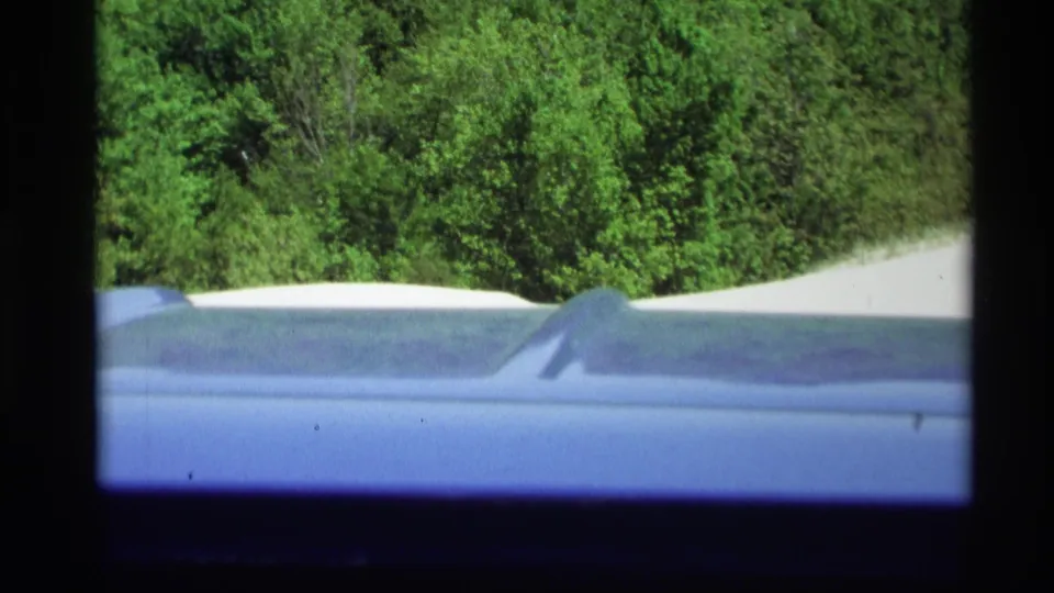 1969 8mm view from a moving car near Holland, Michigan, showing a blue roofline, roadside trees, and sandy dune terrain.
