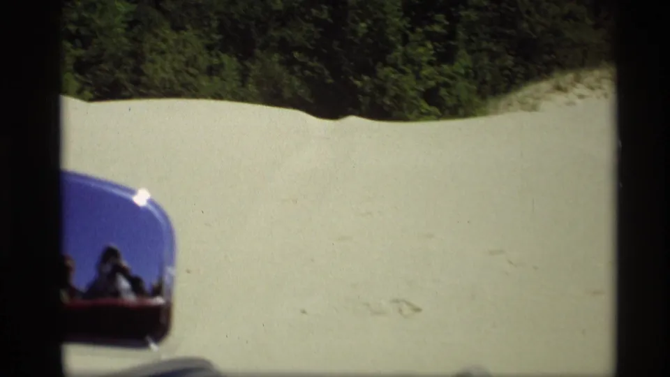 Moving-car view in Holland, Michigan, with a side mirror reflecting passengers while sandy dunes and trees pass outside.