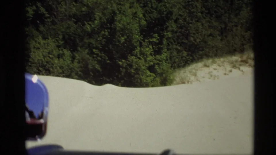 Late-1960s 8mm passenger view from a car near Holland, Michigan, with a partial side mirror, pale sand, and dense green trees.