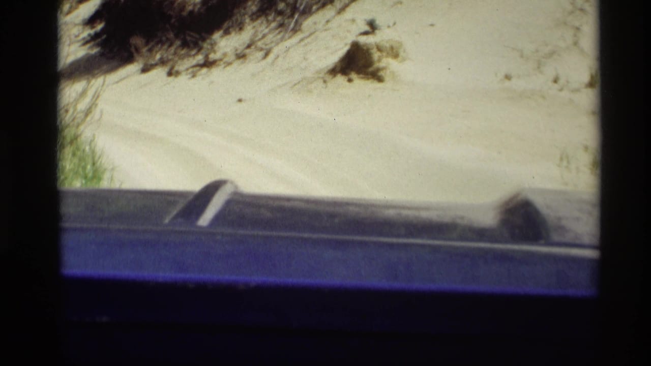 1969 home-movie view near Holland, Michigan, looking past a dark blue vehicle edge toward pale sand and dune vegetation.