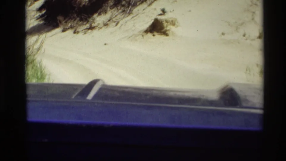 1969 home-movie view near Holland, Michigan, looking past a dark blue vehicle edge toward pale sand and dune vegetation.
