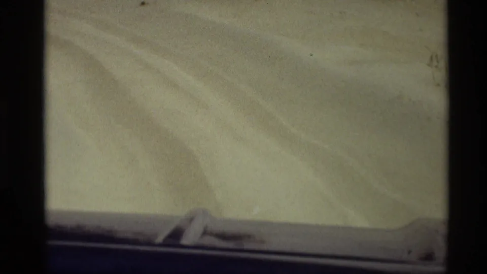 Softly exposed 1969 8mm travel frame showing pale dune sand and curving tracks beyond the blue vehicle edge.