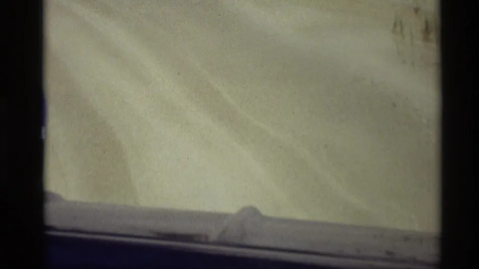 Home-movie frame from a blue vehicle crossing sandy dunes, with faint tire tracks and a dark film border visible.