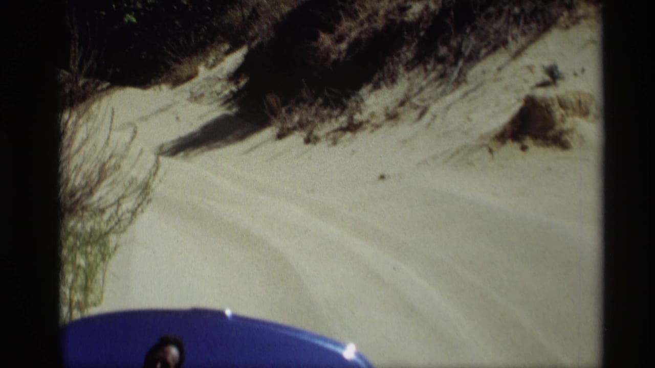 1969 8mm frame from Holland, Michigan showing pale sand, dune grass, and a partial blue foreground object with a person barely visible.