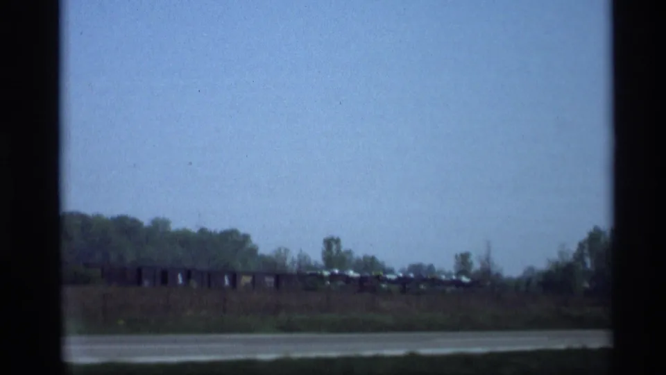 Distant roadside or water-edge view in Holland, Michigan, with low vegetation, pale sky, and small rail cars or vehicles along the horizon.
