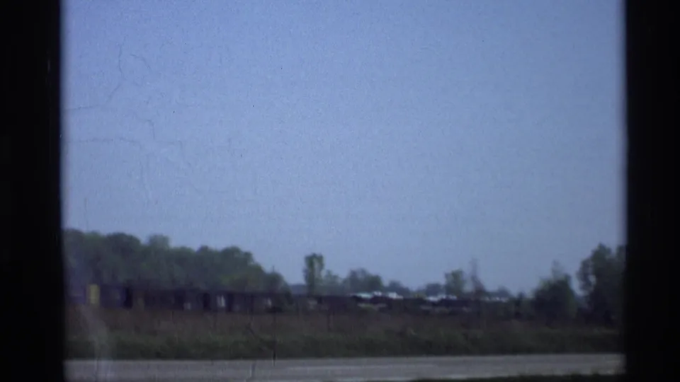 Soft 8mm frame of a rural Holland, Michigan view with open sky, low brush, a road or water edge, and distant rail cars or vehicles.
