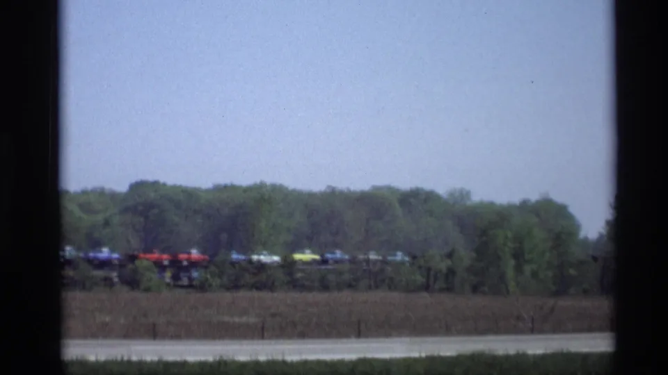 Distant line of colorful vehicles or rail cars in a rural 1969 Holland, Michigan scene, with trees and low brush in the background.