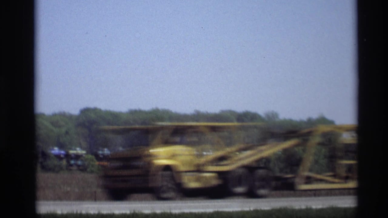 Blurred yellow car carrier passes on a highway with a distant freight train and trees behind it.