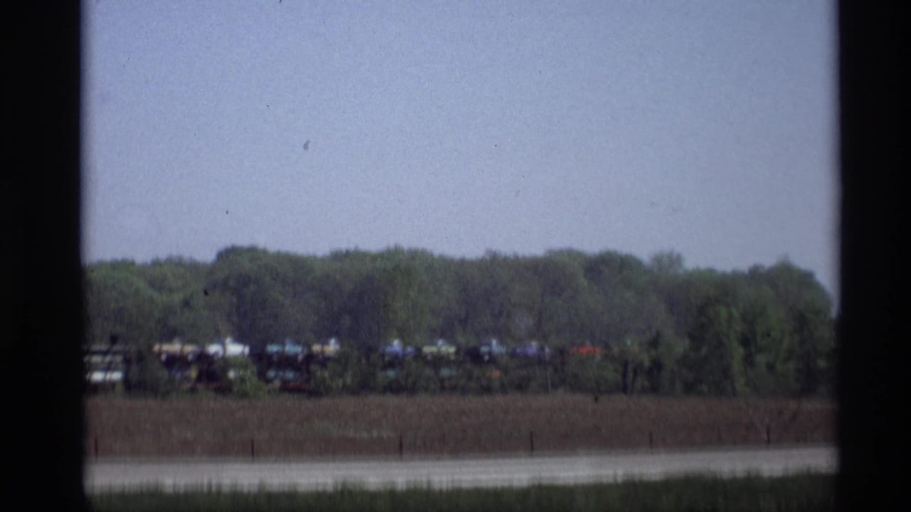 A roadside view in 1969 Holland, Michigan shows a distant freight train beside trees beyond the highway.