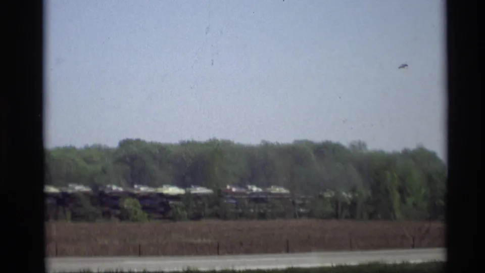 A moving vehicle frames a Holland, Michigan highway scene with distant freight cars, trees, and 1969 home-movie softness.