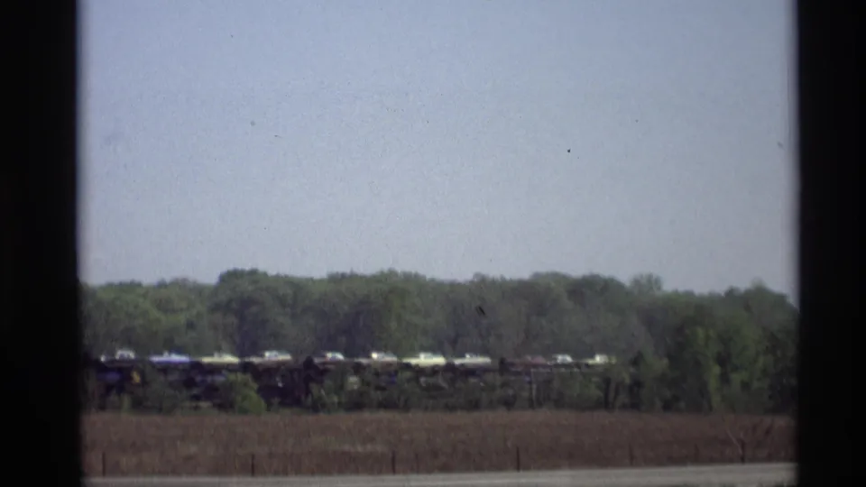 1969 Holland, Michigan home-movie frame showing a distant freight train beyond a field, viewed through a dark vertical window divider.