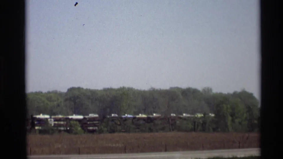 A distant 1969 Holland, Michigan rail scene shows flatcars carrying automobiles, with field, trees, and pale sky in view.