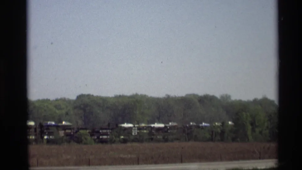 The 8mm frame shows a rural Holland, Michigan view with distant train cars, a field, roadside strip, and soft treeline.