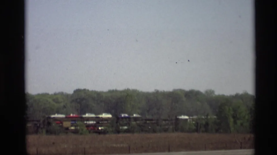 A rural 1969 Holland, Michigan frame shows railcars with automobiles while a small red vehicle blur passes near the road below.
