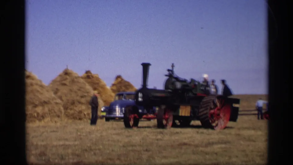 Rural Jordan, Minnesota field scene with a steam tractor, haystacks, blue car, and men standing nearby in 1969 home-movie footage.