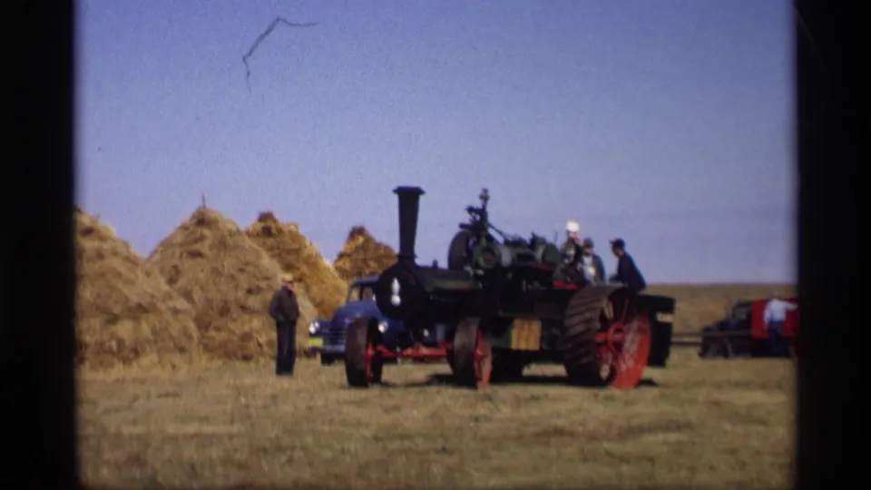 Wide 1969 rural Minnesota home-movie view of a steam tractor beside haystacks, with men and parked cars in the field.