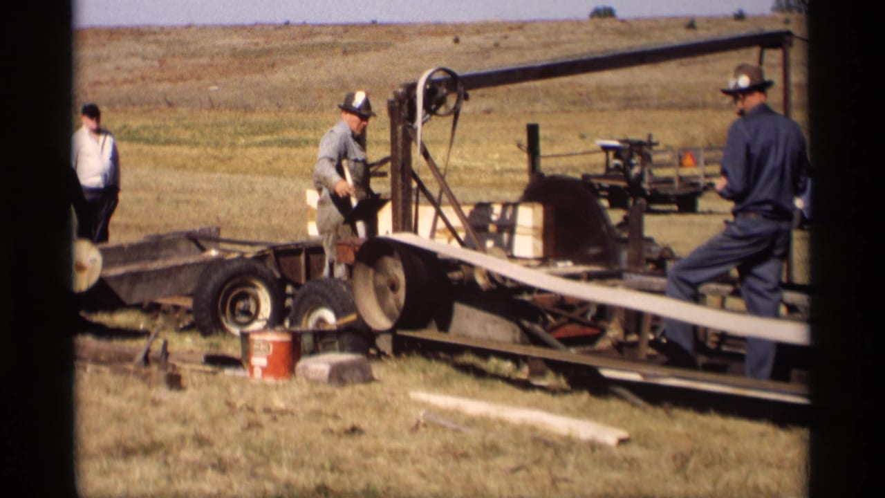 In Jordan, Minnesota in 1969, two men work around a trailer-mounted band saw with a long wooden slat in a dry open field.