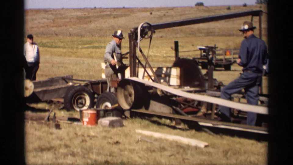 In Jordan, Minnesota in 1969, two men work around a trailer-mounted band saw with a long wooden slat in a dry open field.