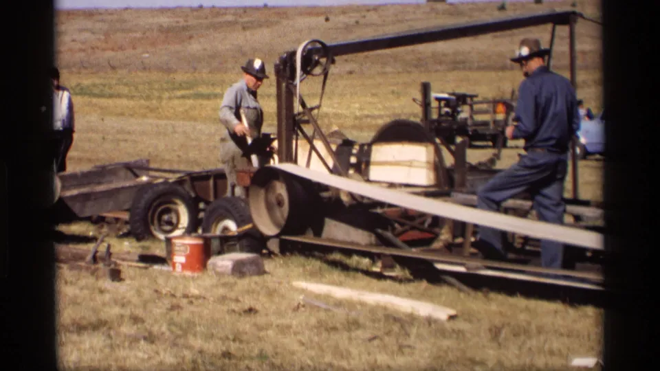 A 1969 rural Minnesota frame shows a man beside a large portable saw and another man standing near the long board feed path.