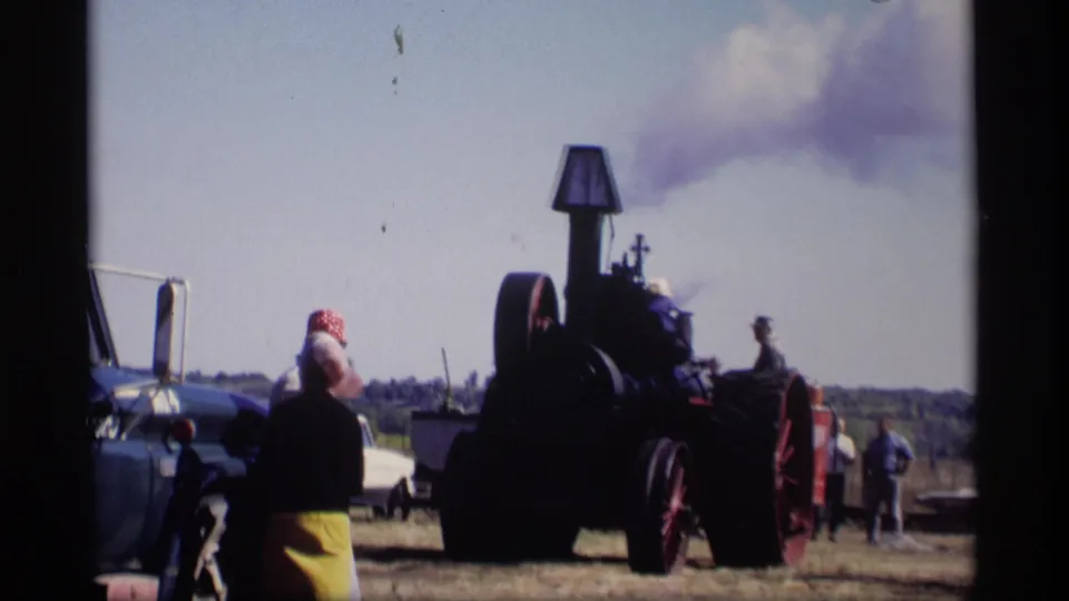 At the 1969 Jordan, Minnesota gathering, spectators watch a dark steam-powered threshing machine with smoke rising nearby.