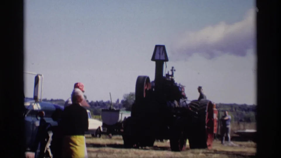 Wide 8mm view of a steam threshing demonstration in Jordan, Minnesota, with spectators standing near the machinery.