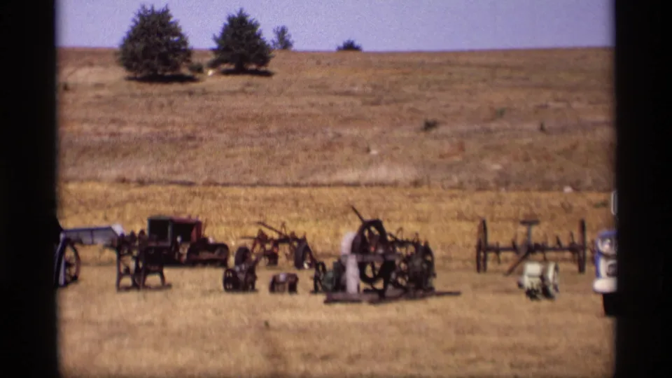 1969 Jordan, Minnesota wide shot of a dry field with antique farm machinery, small tractor-like equipment, fencing, and trees in the background.