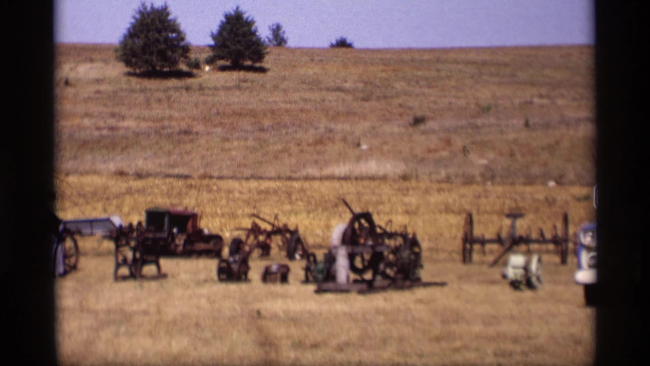 Antique buggies and equipment lined up in a dry field with distant trees and hillside.