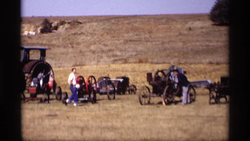 Two adult visitors examine antique buggies and carriages in a rural Jordan, Minnesota field, with dry grass and a hillside behind them.