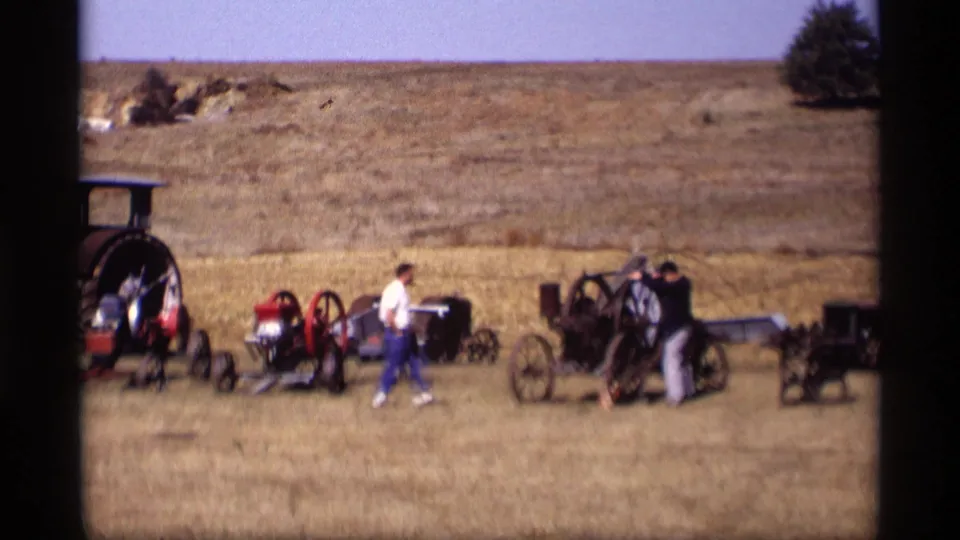 A 1969 home-movie frame shows one adult walking past displayed carriages while another inspects old equipment in the open field.
