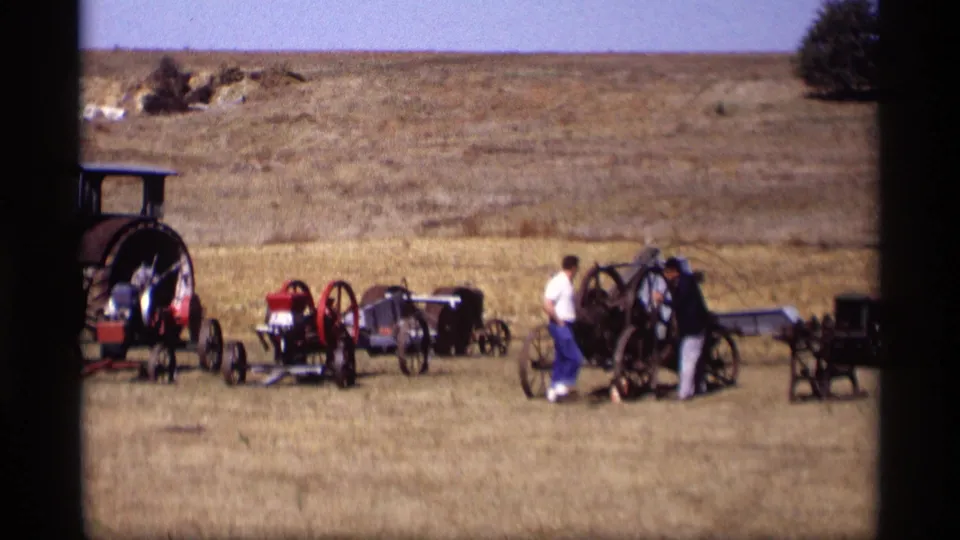 Two adult visitors stand near a large antique wheeled carriage or equipment piece in Jordan, Minnesota, with other buggies arranged nearby.
