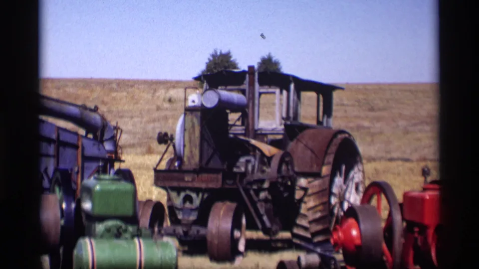 A vintage tractor with large rear wheel stands among farm machines in a Jordan, Minnesota field, filmed on 8mm in 1969.