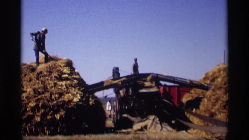 A farm worker stands on a high stack of hay beside the wagon, with additional figures near the equipment in the rural 1969 home-movie scene.